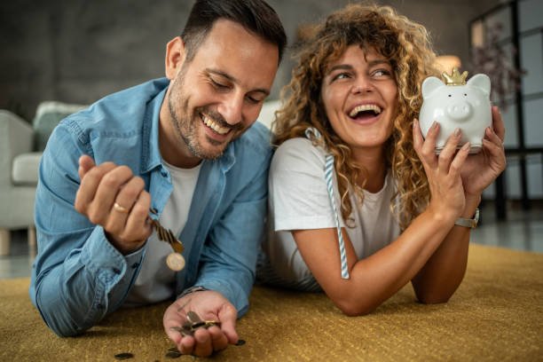 A happy young couple saving money for their next investment, purchase or vacation, they are lying on the living room floor and putting small coins in their small home safe