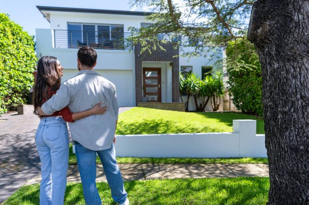 Happy Couple standing in front of their new home. They are both wearing casual clothes and embracing. Rear view. The house is contemporary with a brick facade, driveway, balcony and a green lawn. The front door is also visible.