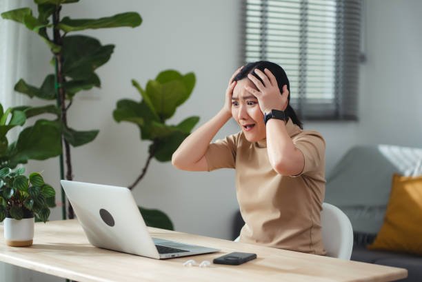 Asian woman sitting at home office thinking about problem with work, shock and feels concerned while looking at laptop.