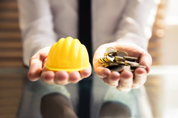 Human Hand Holding Yellow Hardhat And Many Golden Coins Over Desk