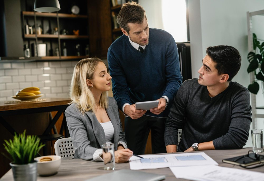 Rear estate agent and young couple going through blueprints and using digital tablet on a meeting 
